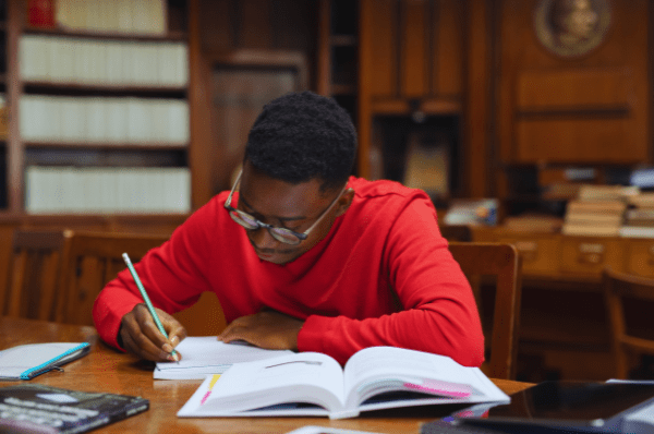 Un étudiant effectue des travaux scolaires dans un bibliothèque. Il porte des lunettes et un chandail rouge, et ses livres et sa tablette électronique sont disposés sur le bureau devant lui. Il écrit dans un cahier à l'aide d'un crayon à mine. L'arrière-plan présente un mur en bois de la bibliothèque, dans lequel plusieurs livres sont exposés.