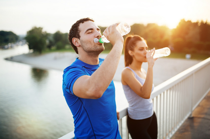 Deux personnes, un homme et une femme, sont habillés avec des vêtements de sport et sont arrêtés sur un pont traversant un rivière. Ils se reposent et boivent de l'eau dans leurs bouteilles respectives. L'arrière-plan est composé de la rivière, d'une forêt, d'une plage et d'un coucher du soleil.