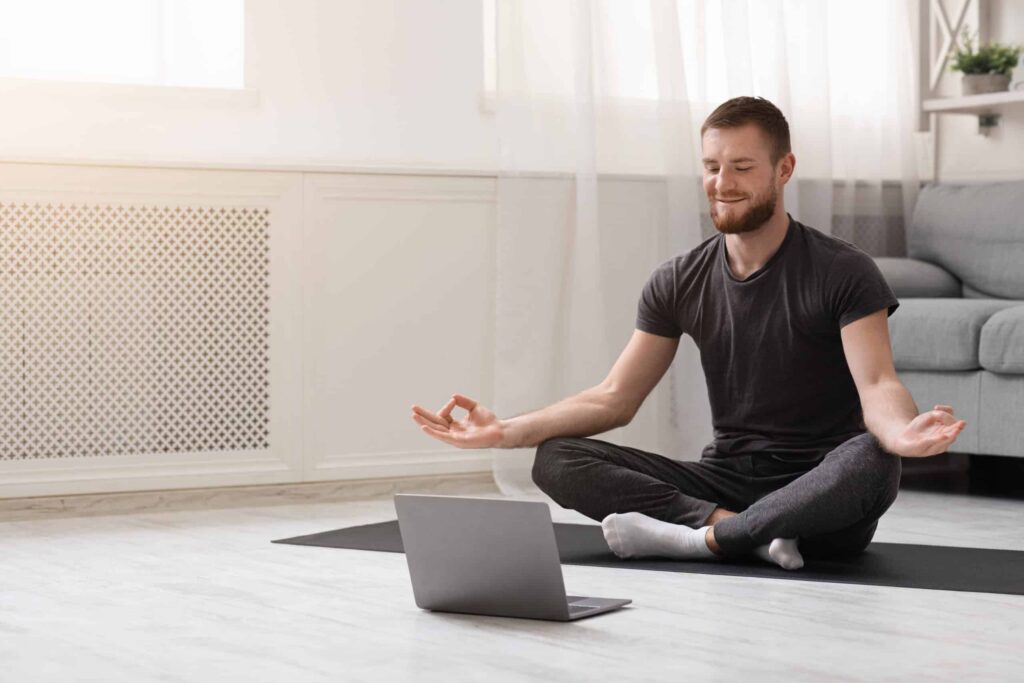 Un homme médite dans son salon. Il est assis sur un tapis de yoga noir au milieu du salon, composé d'un divan gris et de rideaux blancs. Il regarde son ordinateur positionné par terre et exécute la position de méditation de base, soit que ses pouces et ses majeurs respectifs se touchent, ses avant-bras reposent sur ses genoux et ses jambes sont croisées devant lui.