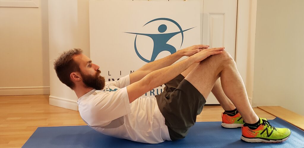 Un homme barbu effectue un redressement assis. Il est dans un environnement intérieur et est assis sur un tapis de yoga bleu. Il effectue le mouvement de redressement de son dos en même temps d'aller porter ses mains aux niveau de ses genoux, dans le but de stimuler et de travailler les muscles abdominaux.