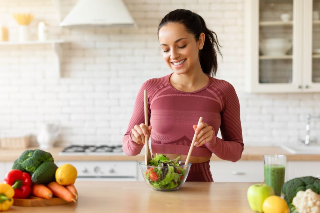 Une femme souriante et habillée en vêtements de sport rouges prépare une salade dans un plat transparent à l'aide de deux spatules en bois. Elle est située dans une cuisine de style moderne, et sur le comptoir en bois franc où elle cuisine se trouve également des poivrons, des brocolis, un avocat, un citron, des carottes, des choux fleurs, une pomme verte et un frappé aux fruits.