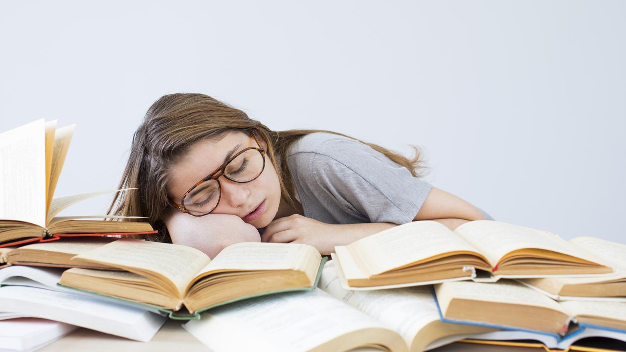 Une jeune femme portant des lunettes est endormie sur une pile de livres ouverts, probablement en plein travail ou étude. La personne, au centre de l'image, repose sa tête sur son bras, entourée de livres ouverts, symbolisant la surcharge de travail ou l'excès d'études. L'arrière-plan est minimaliste et clair.
