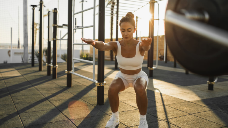 Une femme habillée avec des vêtements de sport blancs fait des exercices physiques en extérieur dans un espace de fitness en plein air, un terrain aménagé avec des équipements sportifs, sous la lumière naturelle du soleil couchant. La femme effectue un squat avec les bras tendus vers l'avant.