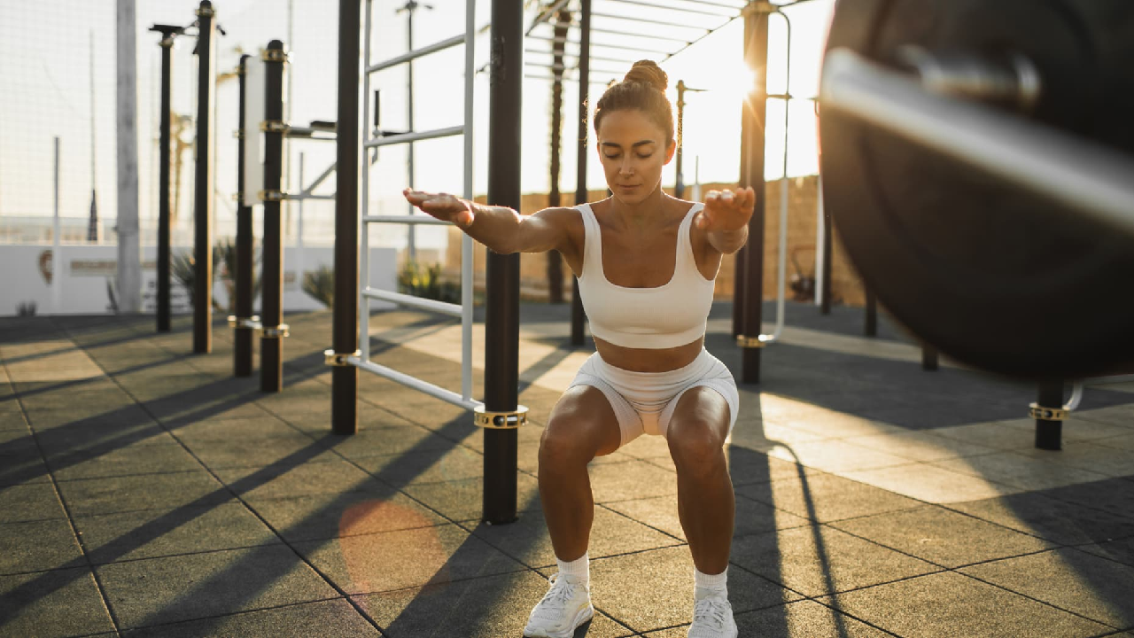 Une femme habillée avec des vêtements de sport blancs fait des exercices physiques en extérieur dans un espace de fitness en plein air, un terrain aménagé avec des équipements sportifs, sous la lumière naturelle du soleil couchant. La femme effectue un squat avec les bras tendus vers l'avant.