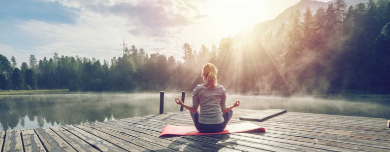 Une femme aux cheveux attachés portant un chandail gris est assise sur un tapis de yoga rouge sur un quai en bois sur le bord d'un lac. Elle est de dos à la caméra, elle regarde au loin vers le lac et vers la forêt. Un pylône électrique est situé dans l'arrière-plan gauche et une montagne est située dans l'arrière-plan droit. Il y a un léger brouillard à la surface de l'eau et le soleil illumine le visage de la femme. Celle-ci est en position de méditation. Elle forme un cercle avec chacune de ses mains, où ses majors et ses pouces respectifs se touchent. Ses jambes sont croisées devant elle et ses coudes touchent ses genoux du même côté.