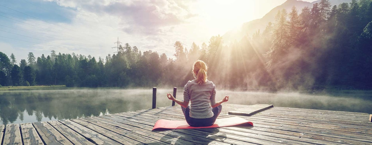 Une femme aux cheveux attachés portant un chandail gris est assise sur un tapis de yoga rouge sur un quai en bois sur le bord d'un lac. Elle est de dos à la caméra, elle regarde au loin vers le lac et vers la forêt. Un pylône électrique est situé dans l'arrière-plan gauche et une montagne est située dans l'arrière-plan droit. Il y a un léger brouillard à la surface de l'eau et le soleil illumine le visage de la femme. Celle-ci est en position de méditation. Elle forme un cercle avec chacune de ses mains, où ses majors et ses pouces respectifs se touchent. Ses jambes sont croisées devant elle et ses coudes touchent ses genoux du même côté.