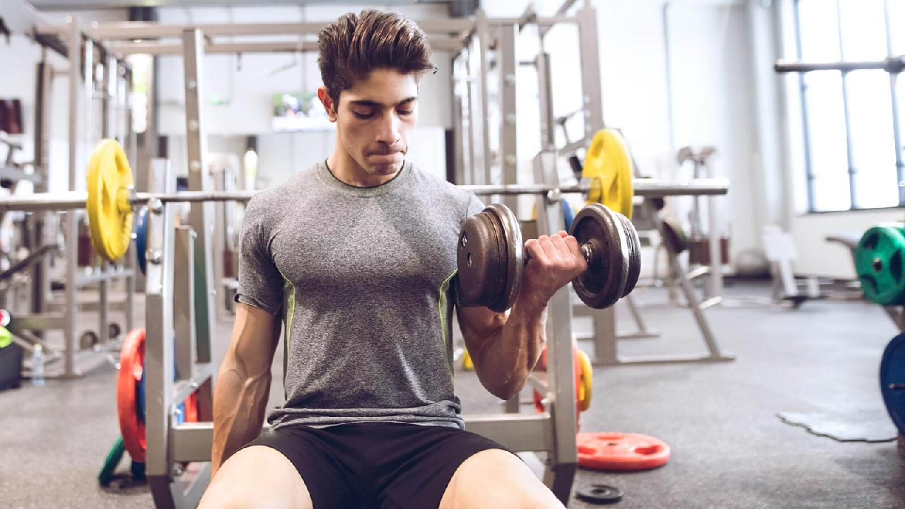 Un jeune homme à la chevelure volumineuse et de couleur brune, et portant un chandail de sport gris s'entraîne seul dans une salle de musculation. Il soulève du bras gauche un haltère de façon à exécuter un exercice travaillant le biceps. Ses veines sont visibles sur ses deux bras. Son visage a une expression d'effort, l'exercice qu'il effectue requiert une certaine force. Dans l'arrière-plan flou se retrouvent diverses installations de musculation, dont une plus visible servant aux exercices de développé couché. Des plaques de poids sont étendues sur le sol.
