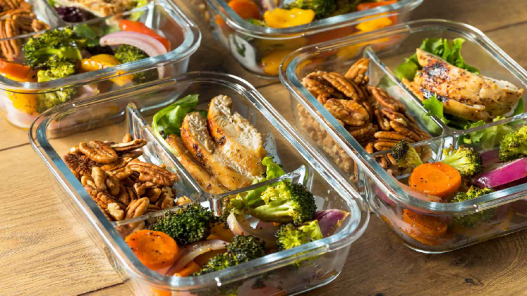 Quatre repas identiques et déjà préparés (prêts à manger) sont positionnés dans des plats de vitres sans couvercle. Ceux-ci contiennent tous des morceaux de poulet grillé couchés sur une feuille de laitue dans le compartiment en haut à droite, des noix de pécan dans le compartiment en haut à gauche et des légumes grillés dans le compartiment du bas, tels que des carottes tranchées, des brocolis et des morceaux d'oignons. Les plats de vitres sont disposés sur une table en bois franc.