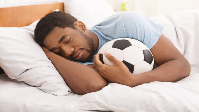 Un homme portant un chandail bleu pâle dort paisiblement, les yeux fermés, sur un lit aux draps blancs et sous une couverture blanche. Sa main droite est posée sous sa tête et sa main gauche tient contre torse un ballon de soccer blanc et noir. Il est positionné de façon à faire face au côté du lit le plus près de lui et fait face à la caméra.